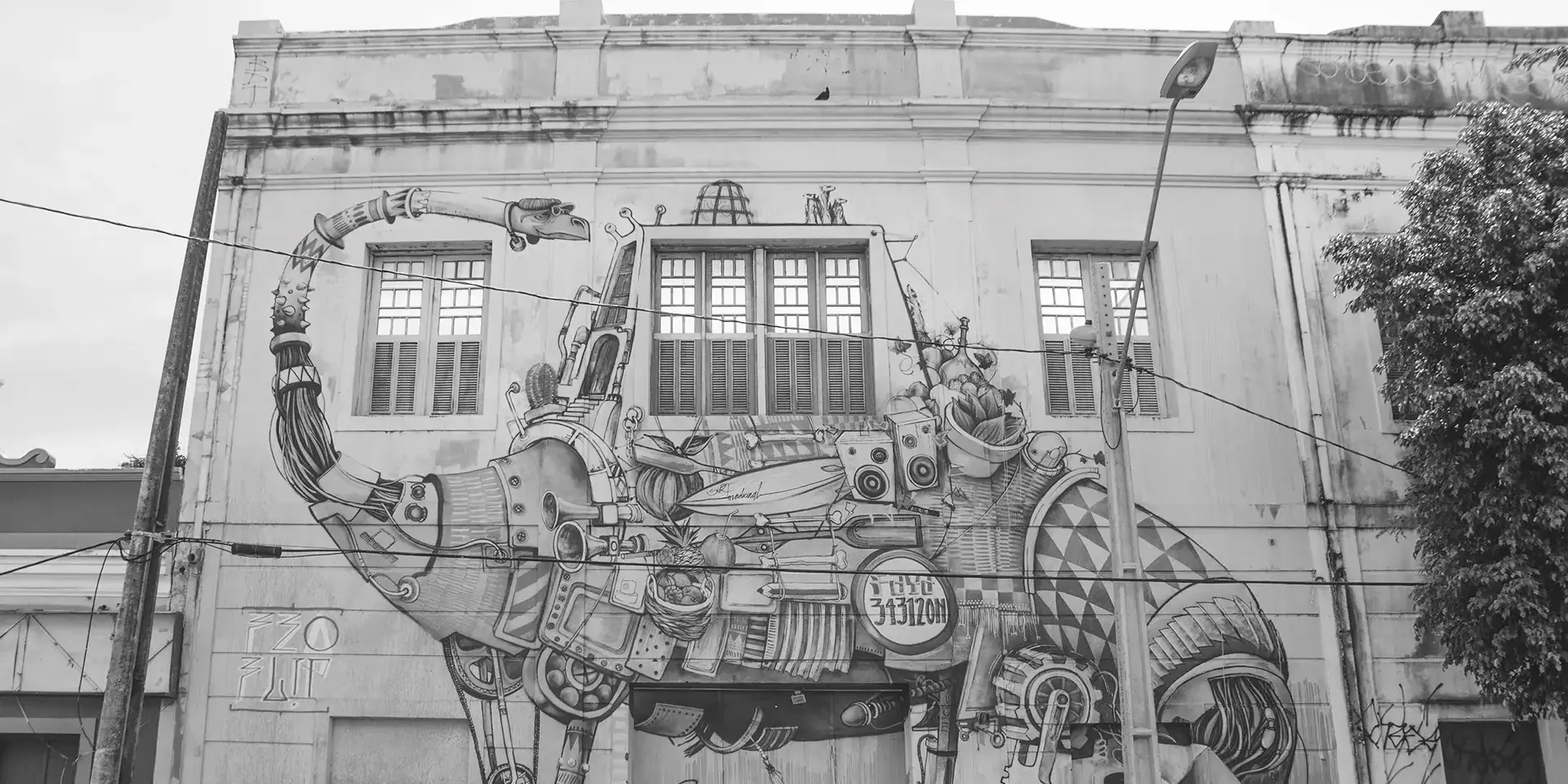 Grayscale photograph of a large mural on the facade of an old building with a futuristic dinosaur with various accessories being observed by a group of people in front of the building