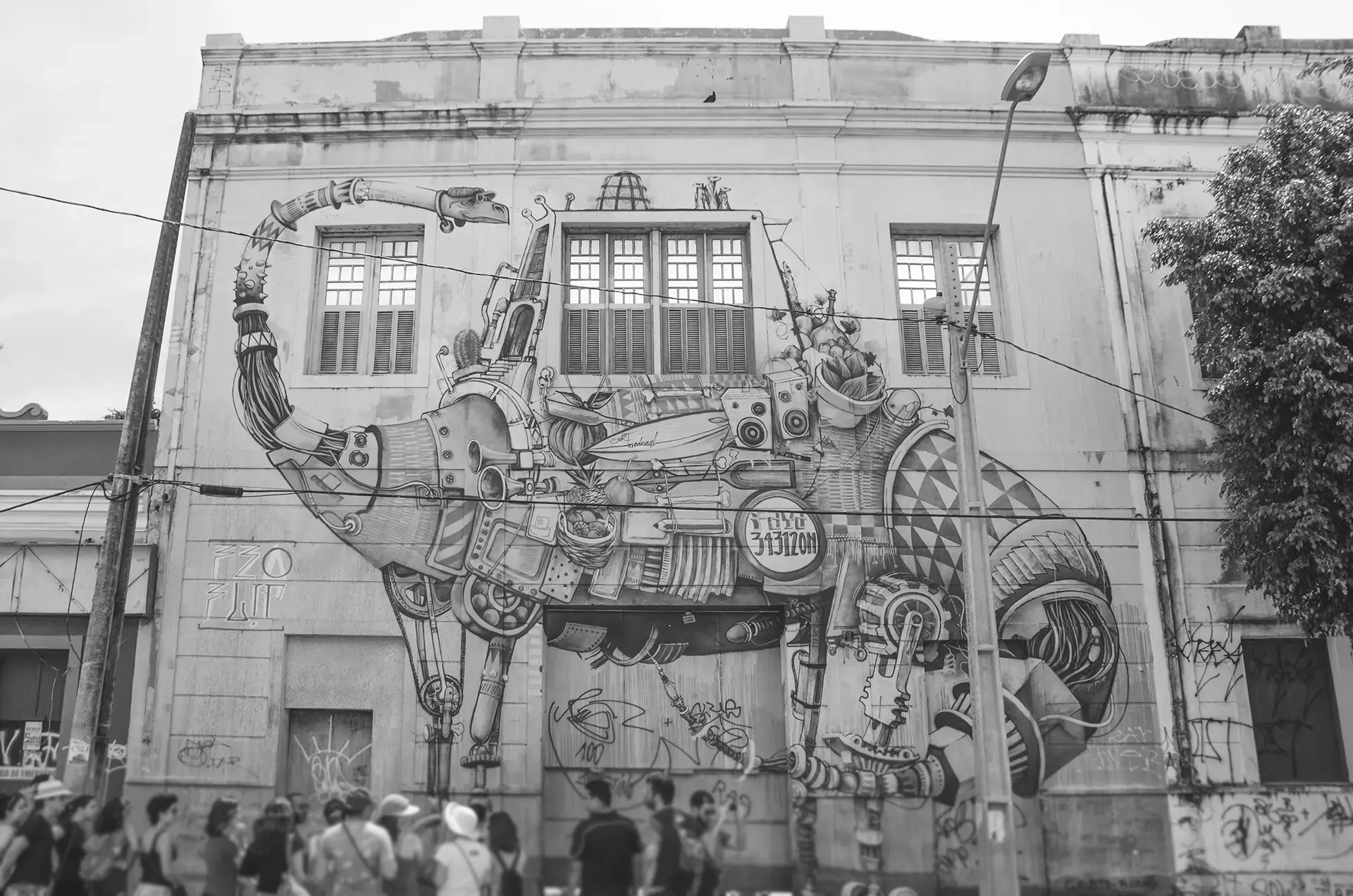 Grayscale photograph of a large mural on the facade of an old building with a futuristic dinosaur with various accessories being observed by a group of people in front of the building