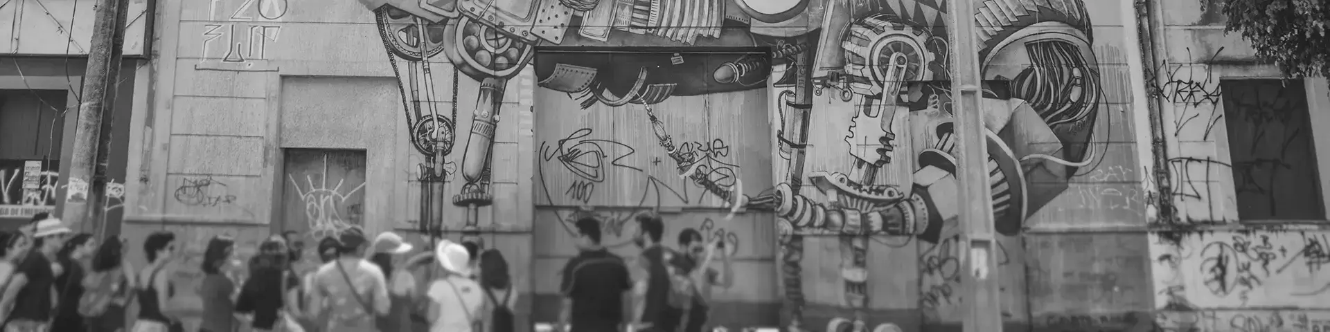 Grayscale photograph of a large mural on the facade of an old building with a futuristic dinosaur with various accessories being observed by a group of people in front of the building