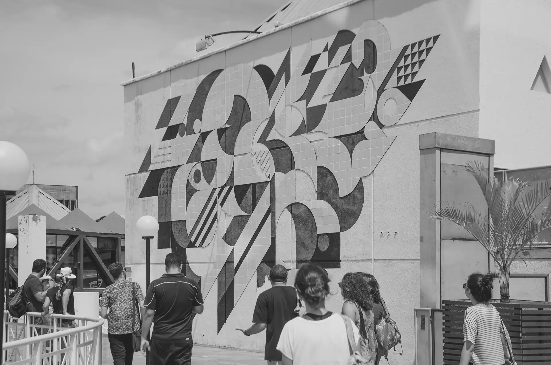 Grayscale photograph of a mural with geometric paintings and tiles with a group of people from behind observing the mural
