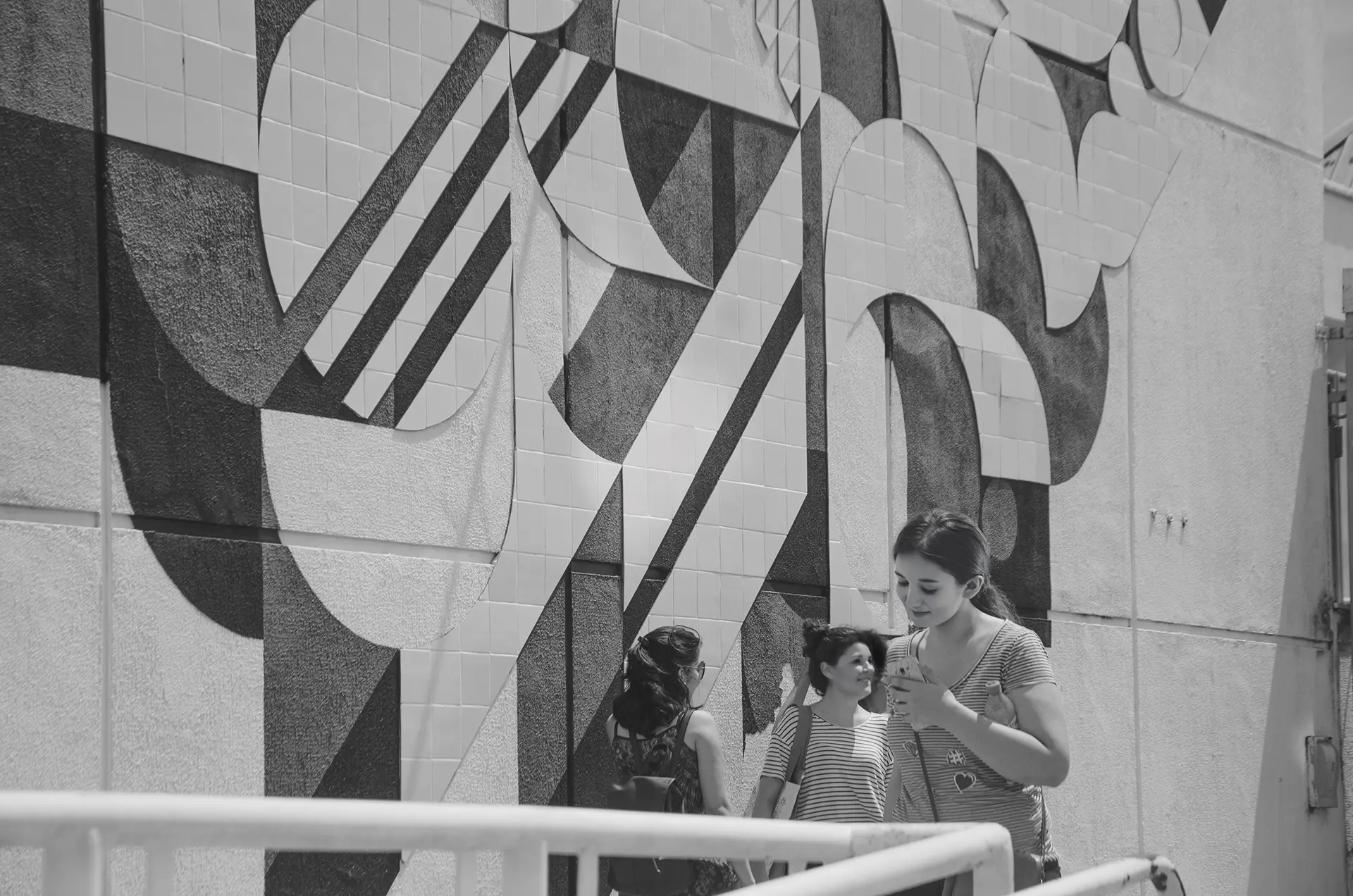 Grayscale photograph of a mural with geometric paintings and tiles with a group of people from behind observing the mural