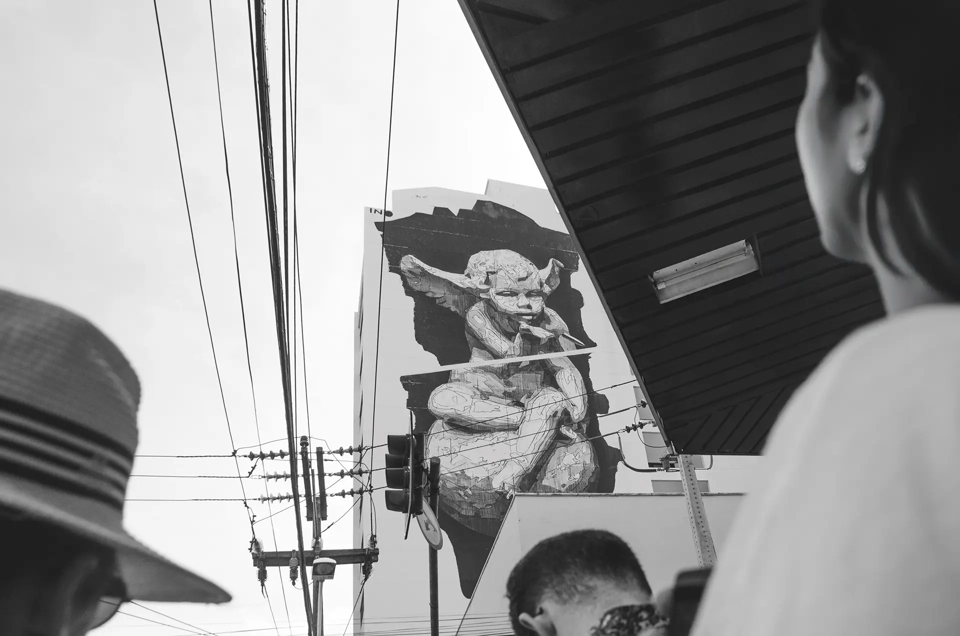 Grayscale photograph of a giant mural on the side of a building with a little angel blowing a bubble in downtown Fortaleza with a man from behind with his head down
