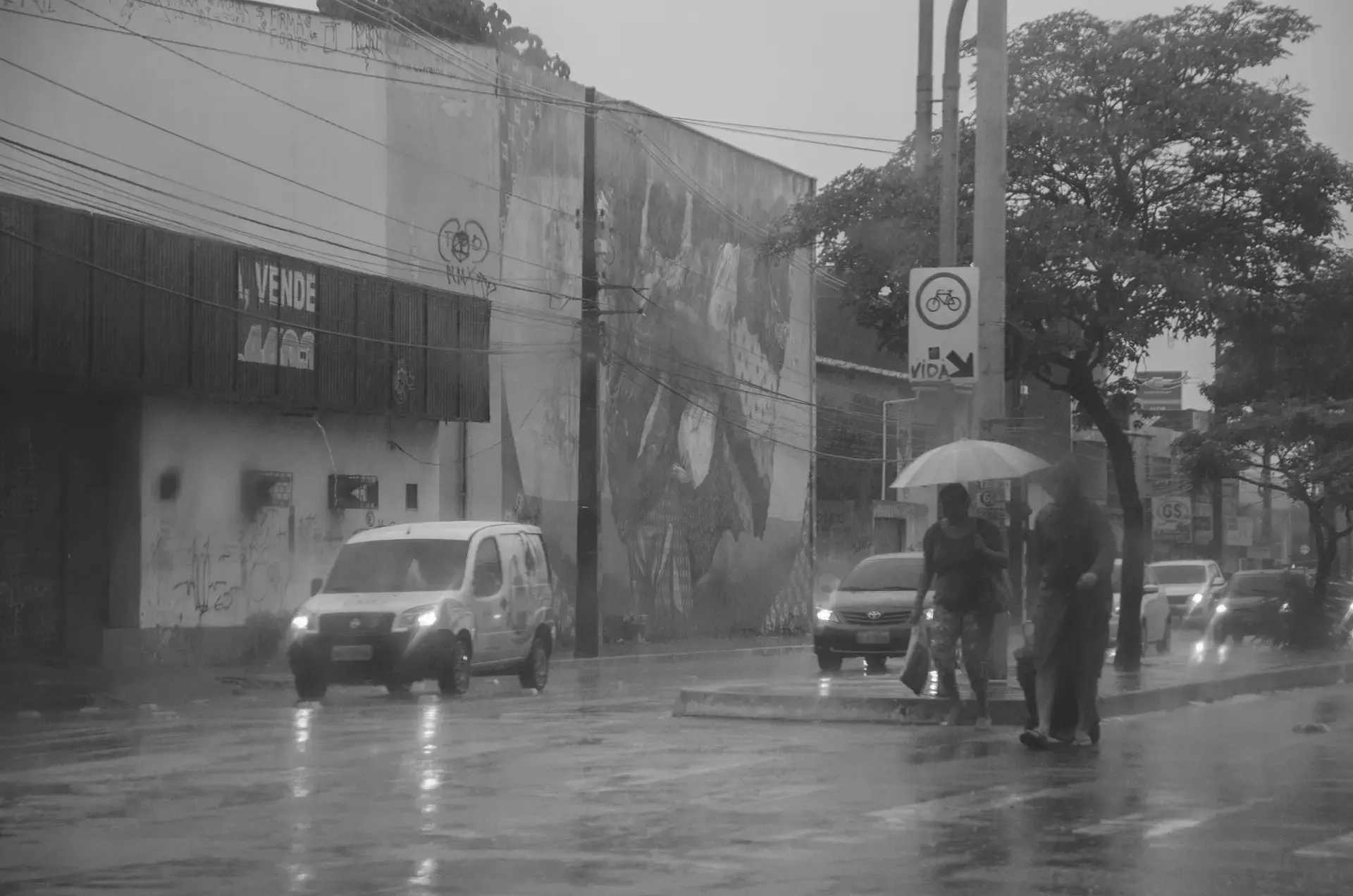Grayscale photograph of a crossing in the Benfica neighborhood in Fortaleza where you can see cars, pedestrians, and graffiti on the wall of a shop in the background