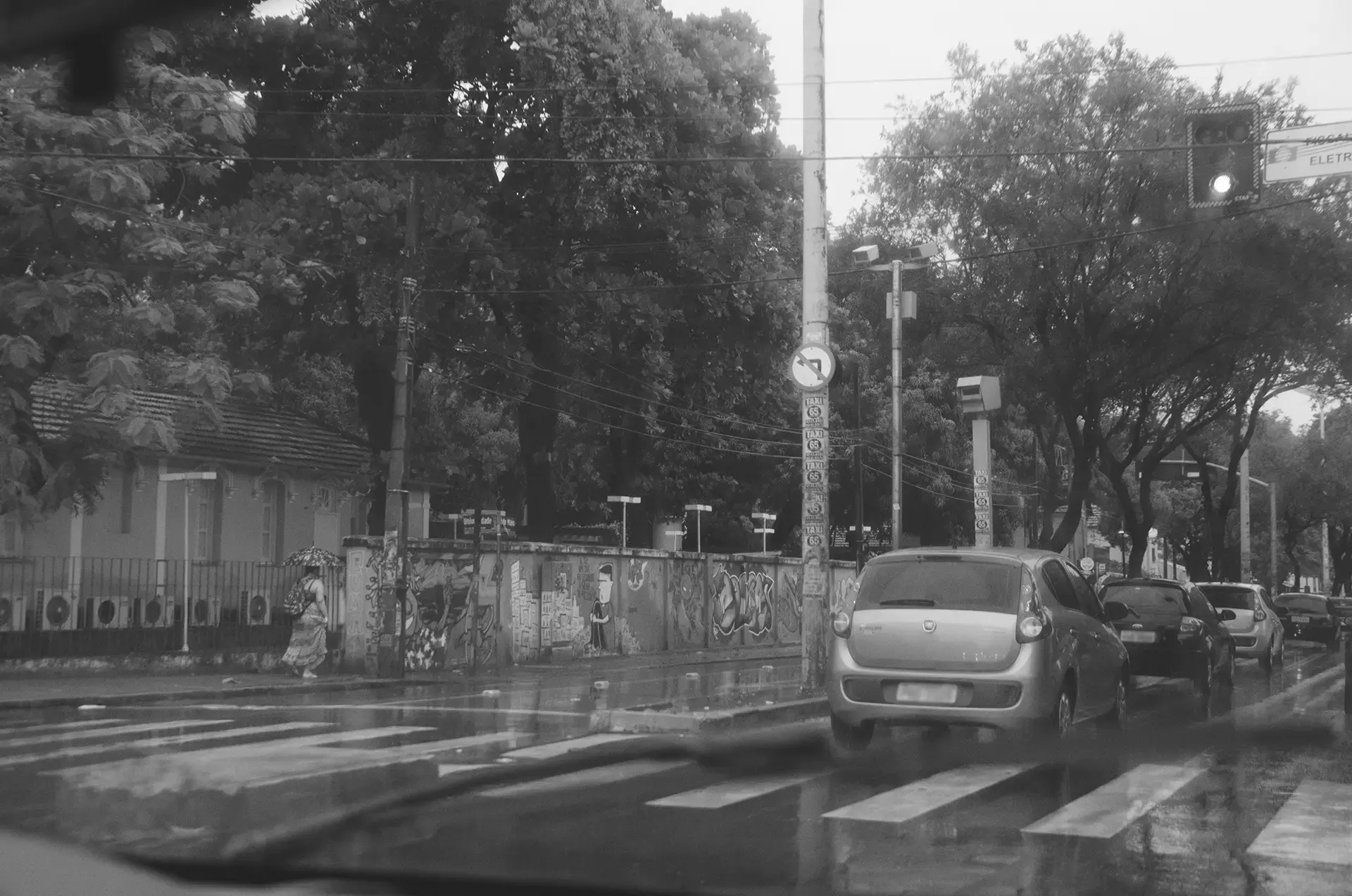Grayscale photograph of cars stopped at a traffic light on Avenida 13 de Maio in Fortaleza during a rainy afternoon where you can see a pedestrian with her umbrella and the walls of the Federal University of Ceará with urban art interventions