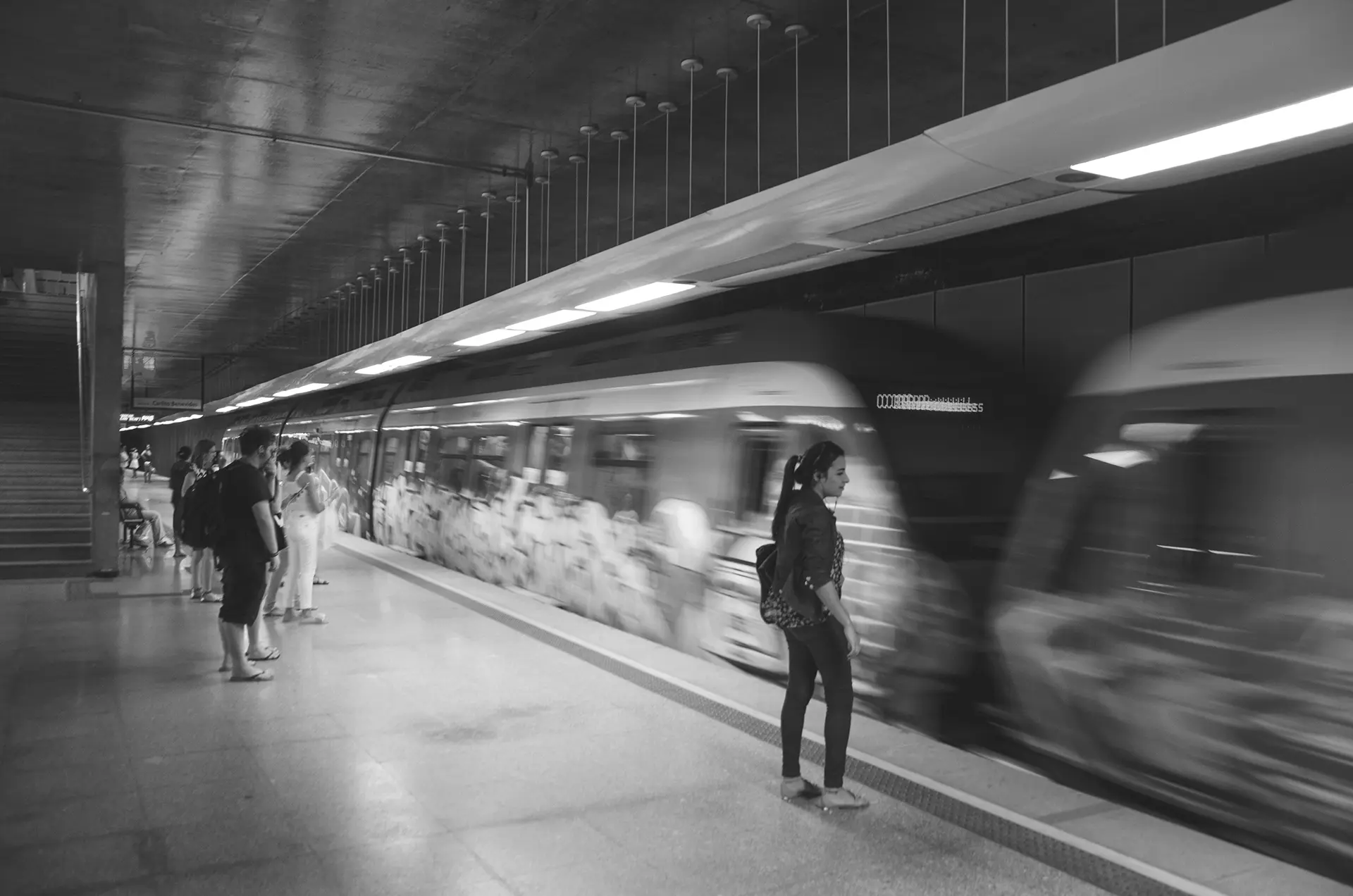 Grayscale photograph of a woman waiting for the train at Benfica Metro Station in Fortaleza, where you can see the graffiti-covered train cars