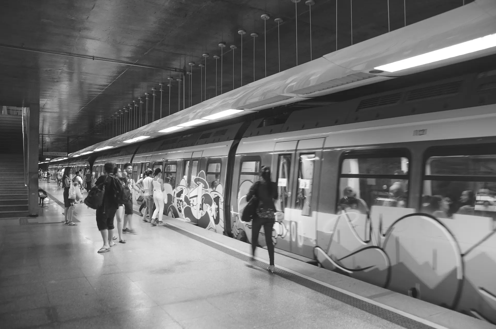 Grayscale photograph of a woman walking to the train door at Benfica Metro Station in Fortaleza, where you can see the graffiti-covered train cars