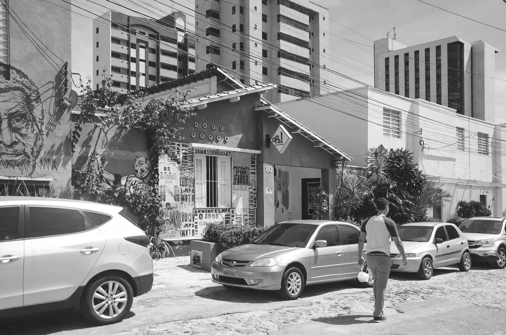 Grayscale photograph of an alley where you can see parked cars, a passerby walking, and walls with urban art interventions