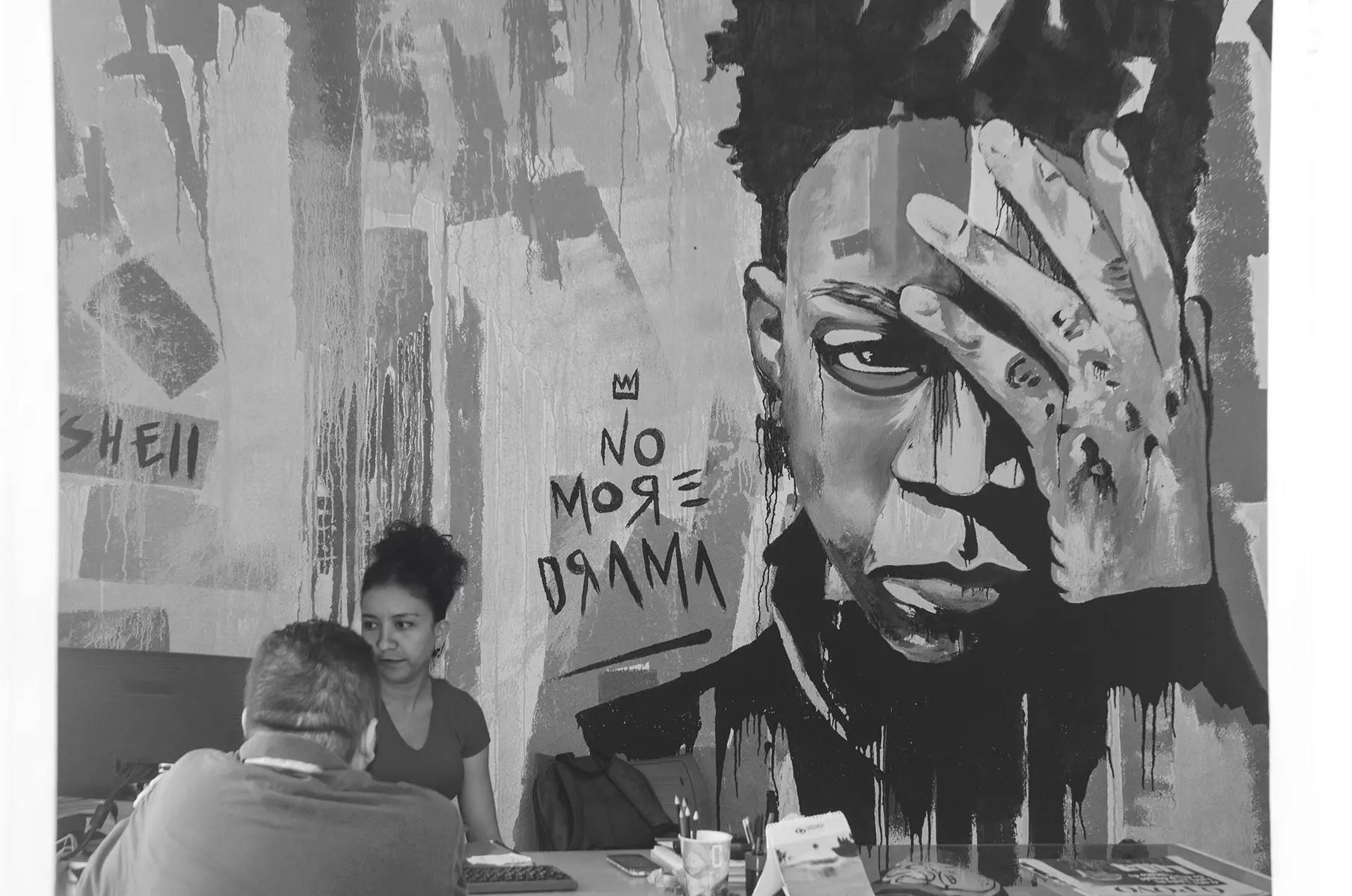 Grayscale photograph of the reception area of a design school with a graffiti mural of Basquiat with his hand on his forehead and the phrase NO MORE DRAMA while a receptionist assists a student