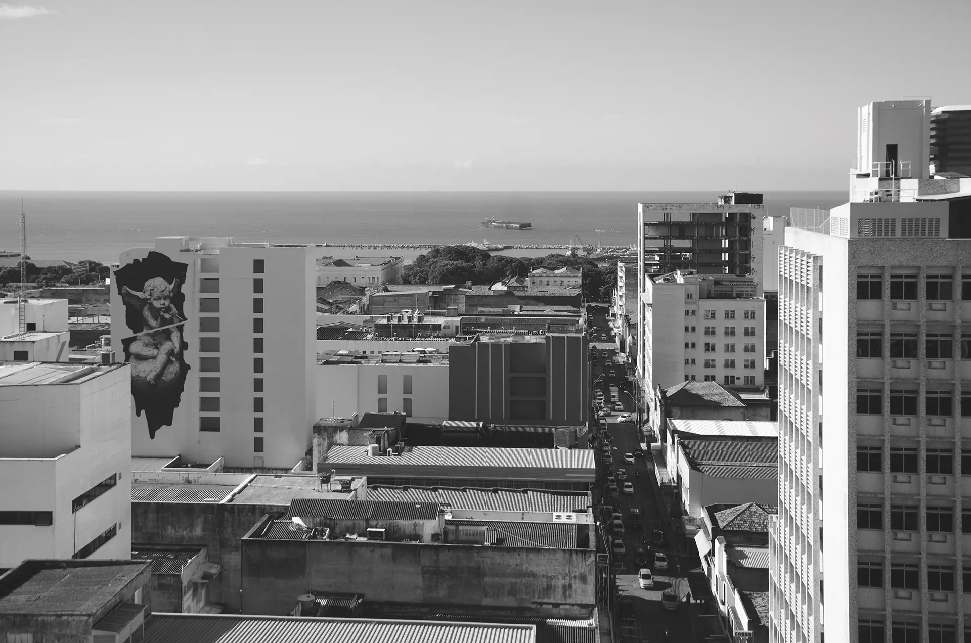 Grayscale photograph in perspective of downtown Fortaleza where you can see several buildings, one of them with a mural of a little angel blowing a bubble, and the city's coastline in the background