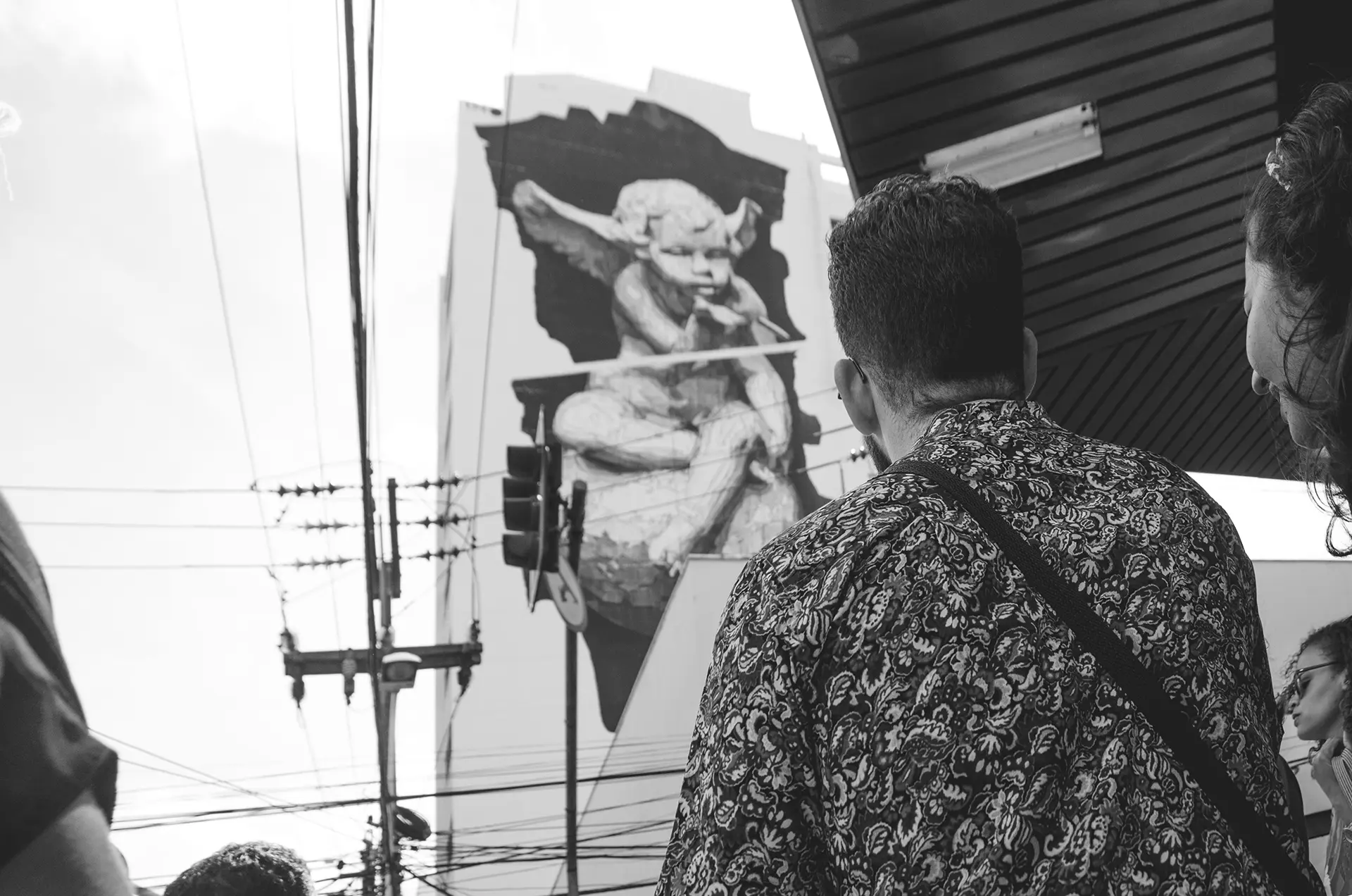 Grayscale photograph of a giant mural on the side of a building with a little angel blowing a bubble in downtown Fortaleza with a man observing it from behind