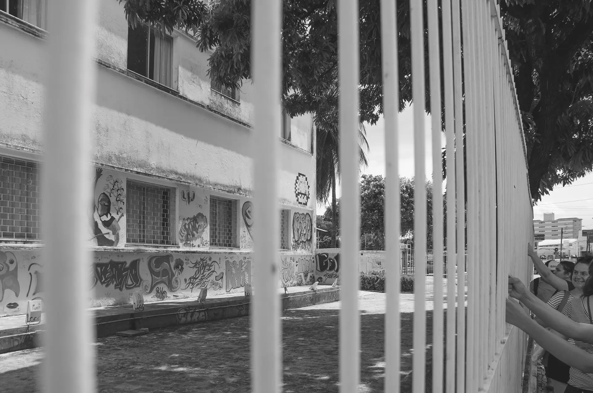 Grayscale photograph in perspective of a fence with people holding it and in the background a building with urban art interventions