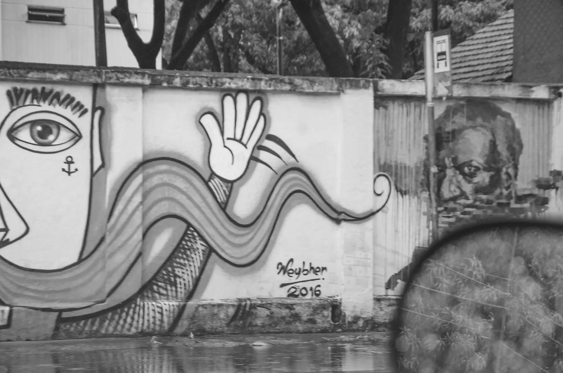 Grayscale photograph of a graffiti wall on Avenida 13 de Maio in Fortaleza taken through a car window where you can see the rearview mirror and in the background one of the murals of a black man looking back intently