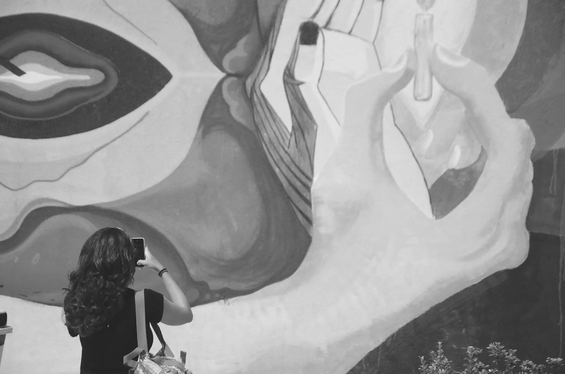 Grayscale photograph of a woman from behind photographing a graffiti mural on the wall of a square in downtown Fortaleza with her cell phone