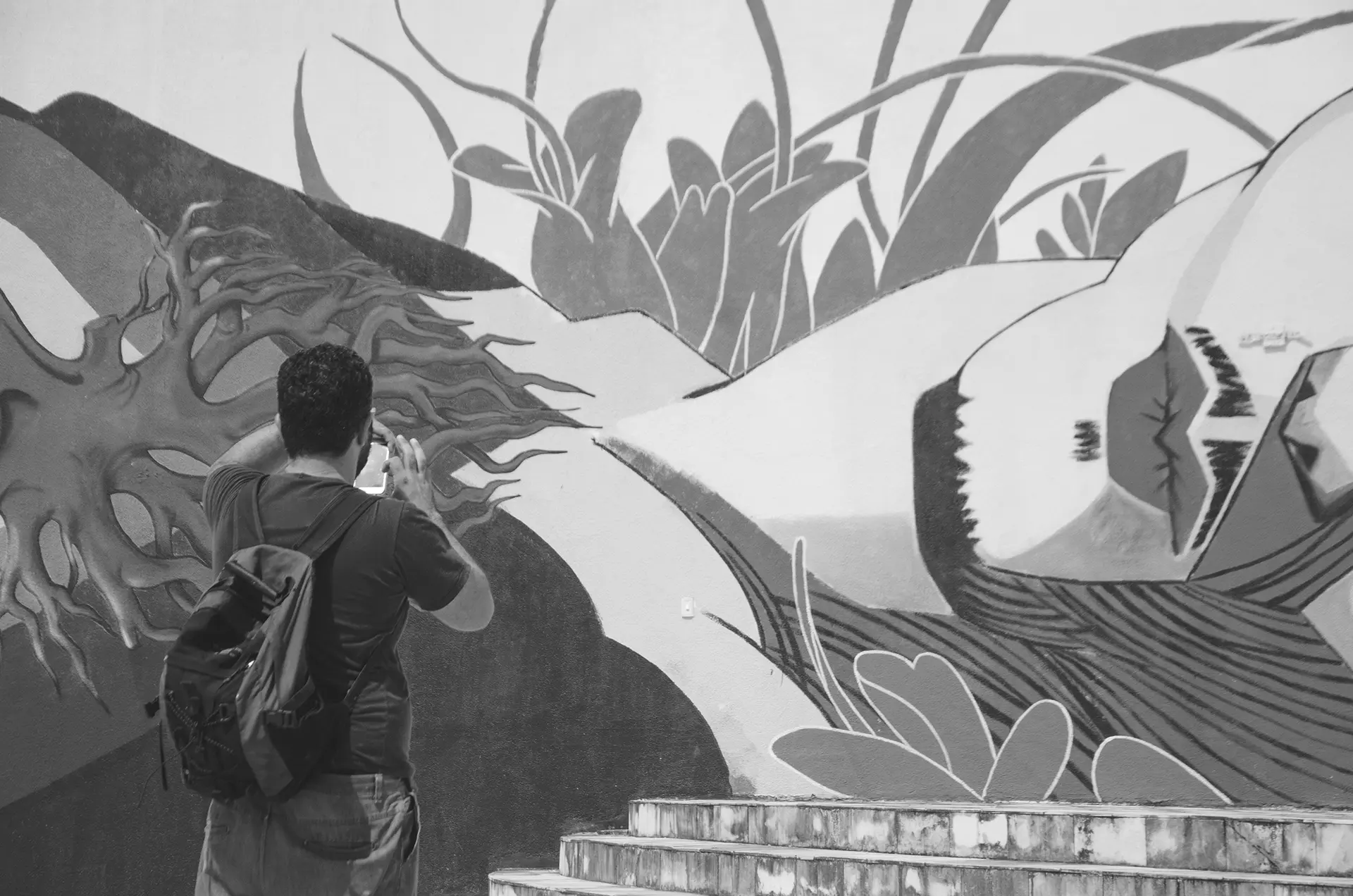 Grayscale photograph of a man from behind photographing a graffiti mural on the wall of a square in downtown Fortaleza with his cell phone