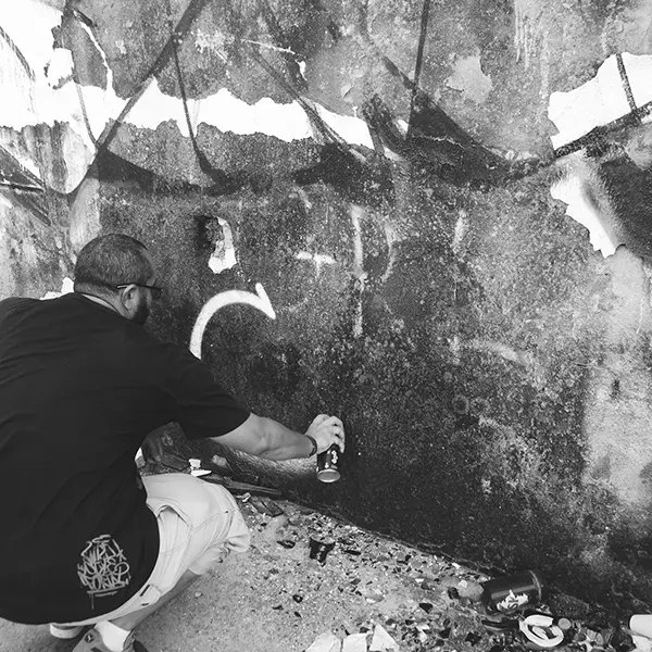 Frame 1 in grayscale from a video of a man squatting with a spray can in his hand while drawing a signature on the wall of an abandoned building