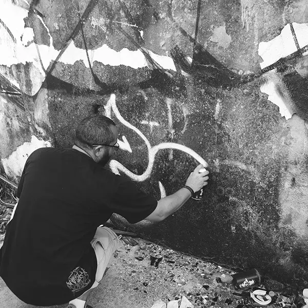 Frame 10 in grayscale from a video of a man squatting with a spray can in his hand while drawing a signature on the wall of an abandoned building