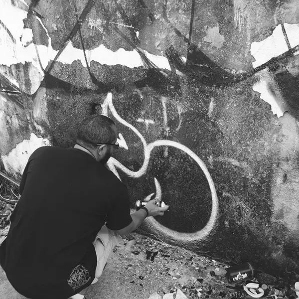 Frame 13 in grayscale from a video of a man squatting with a spray can in his hand while drawing a signature on the wall of an abandoned building
