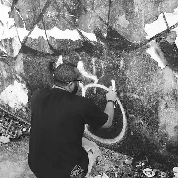 Frame 16 in grayscale from a video of a man squatting with a spray can in his hand while drawing a signature on the wall of an abandoned building