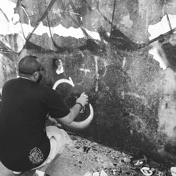 Frame 4 in grayscale from a video of a man squatting with a spray can in his hand while drawing a signature on the wall of an abandoned building