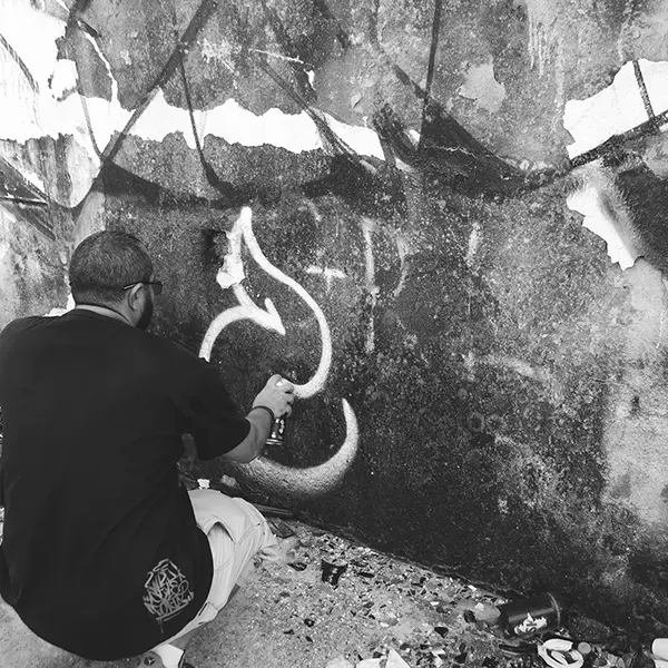 Frame 7 in grayscale from a video of a man squatting with a spray can in his hand while drawing a signature on the wall of an abandoned building
